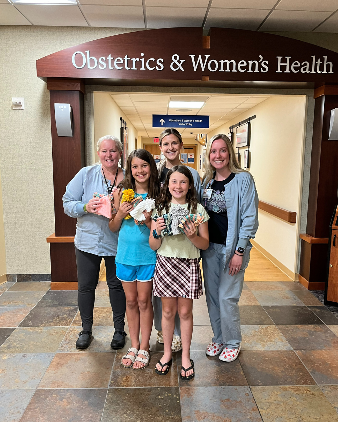 Two 4th grade girls and three medical staff stand in front of the Obstetrics & Women's Health clinic holding up hand-knitted baby hats.